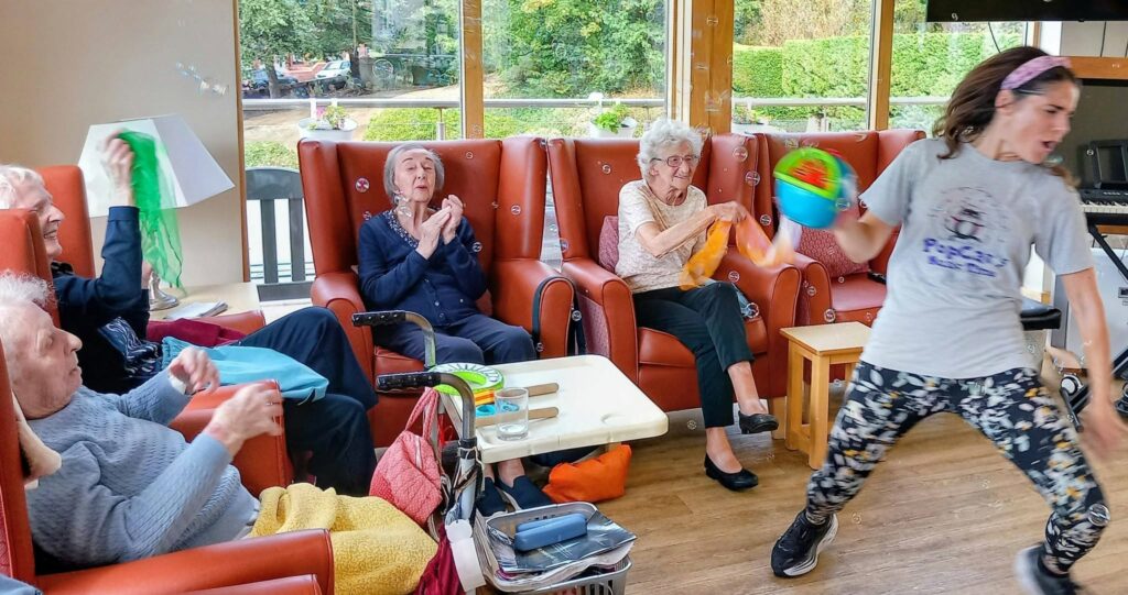 A group of elderly people sit in armchairs, clapping and waving scarves, whilst a young woman in sportswear leads a lively Music and Movement session using a tambourine.