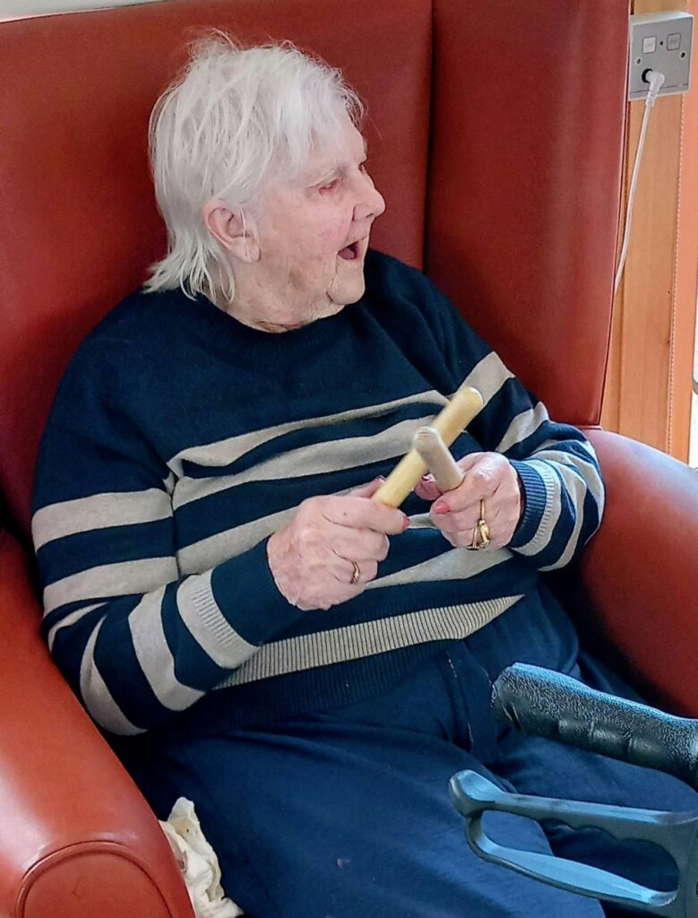 An elderly person with white hair, dressed in a striped jumper, sits in a red armchair holding wooden rhythm sticks, ready for Music and Movement activities.