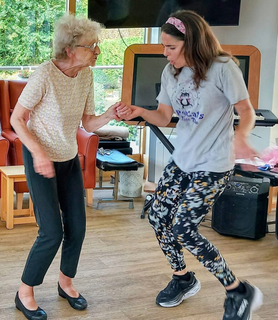 An elderly woman and a younger woman hold hands and dance together indoors, waltzing joyfully among chairs and gym equipment.