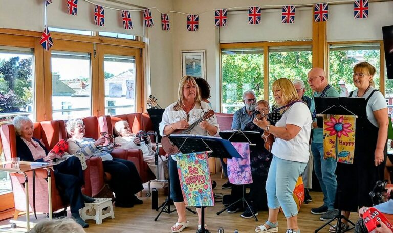 The group N’Ukes perform and sing for an audience in a bright Lear House room adorned with Union Jack flags. Some attendees sit on chairs, smiling and enjoying the show.