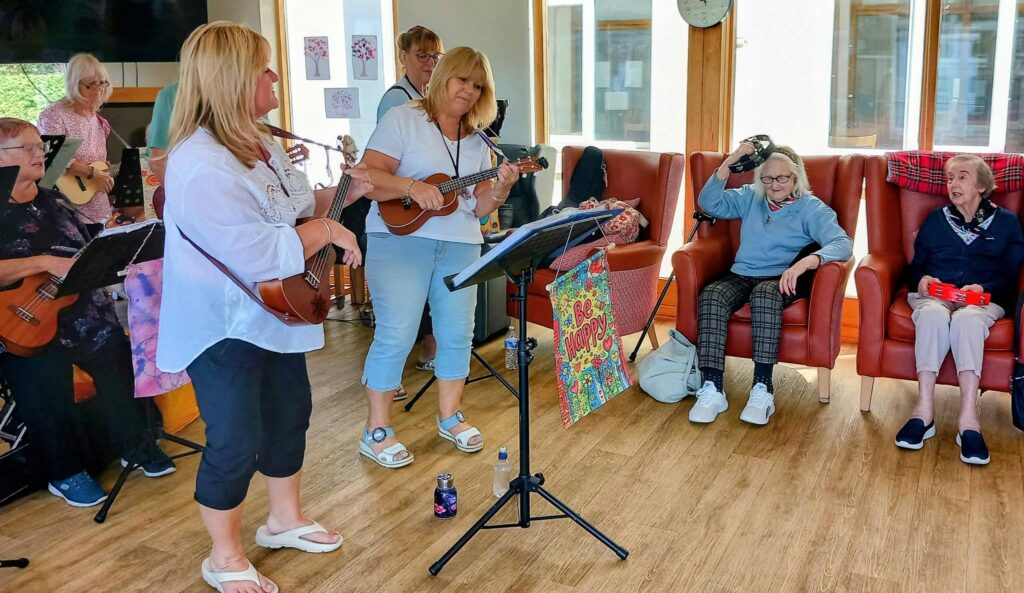 A group of women from 64 N’Ukes play ukuleles in a brightly lit room at Lear House as several elderly women sit on chairs, some holding percussion instruments, enjoying the music.