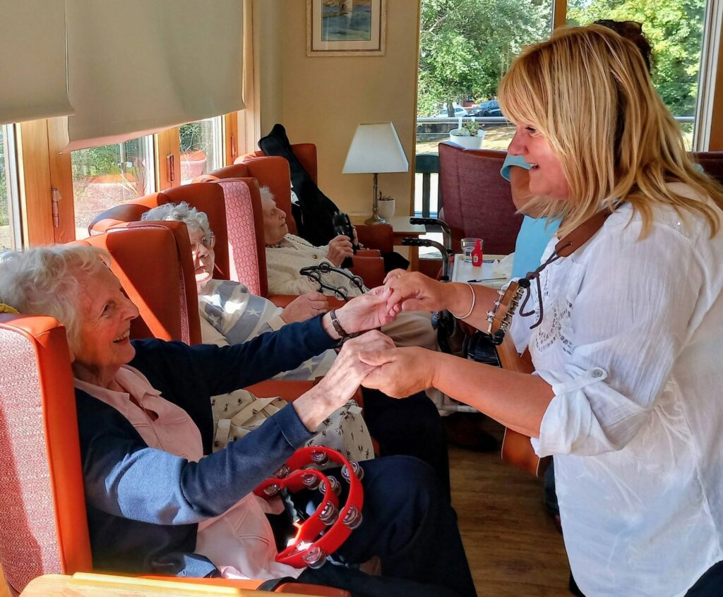 A woman smiles and holds hands with an elderly lady in the lounge at Lear House care home. Sunlight pours in as other residents relax nearby, enjoying music played by the 64 N’Ukes group in the background.