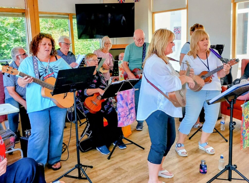 At Lear House, the 64 N’Ukes group fill the brightly lit room with music and laughter as adults play guitars and ukuleles together, reading from music stands while some are standing and others are seated.