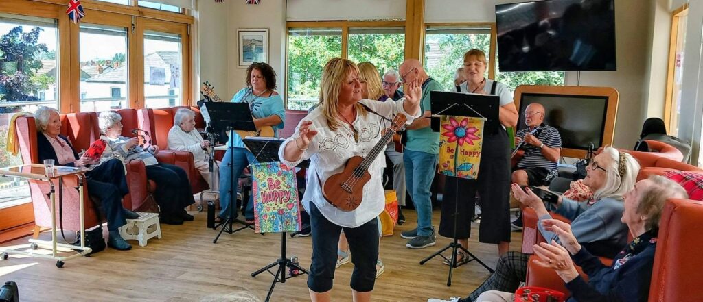The 64 N’Ukes play music for older adults in Lear House’s light-filled common room; the audience clap along as ukulele and guitar music fills the air. Cheerful banners decorate the stands, making for a joyful atmosphere.