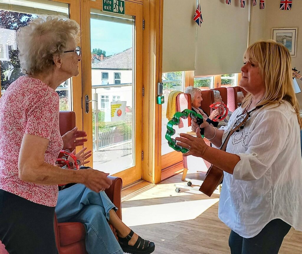 Two women engage with tambourines in a bright room at Lear House, large windows behind them. Others are seated nearby, and Union Jack flags hang from the ceiling, enhancing the musical atmosphere.