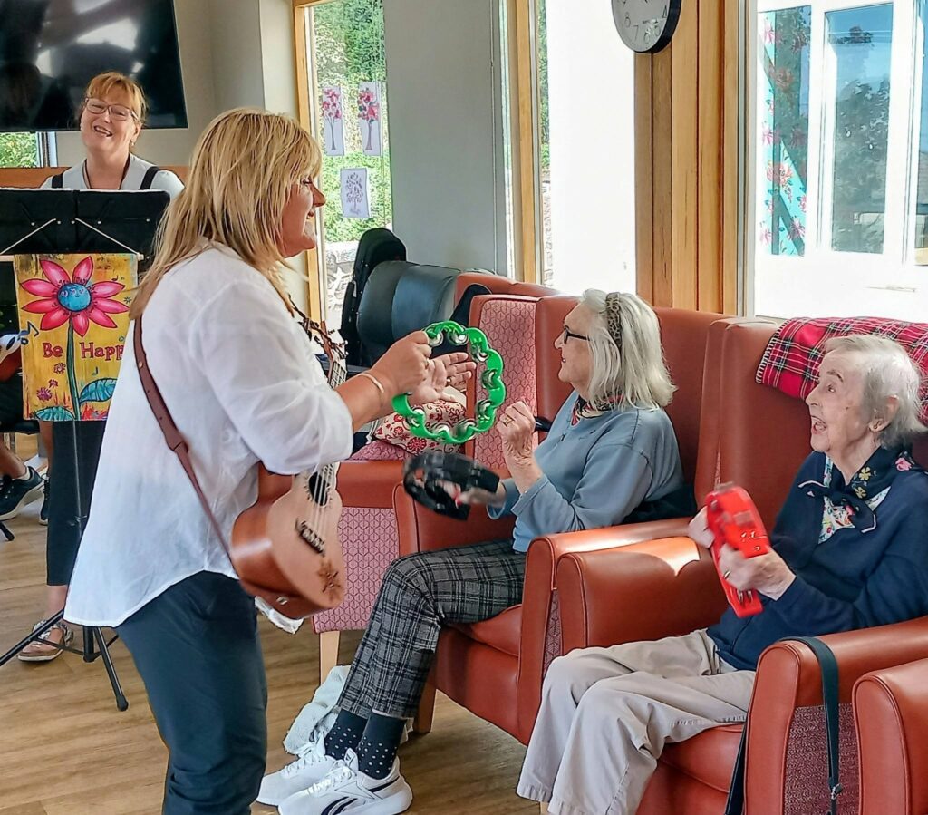 A woman plays guitar and sings whilst two older women seated in armchairs play tambourines at Lear House. Another musician stands nearby, filling the bright, colourful room—perhaps a lively session by 64 N’Ukes.