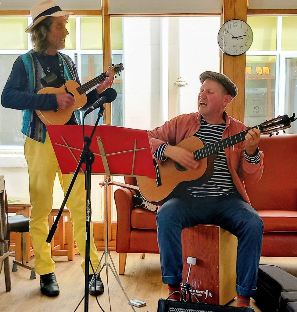Andrew and Marko play music indoors; one stands with a ukulele, the other sits on a cajón playing guitar. Both wear hats as they entertain during their visits, with a clock and large windows in the background.