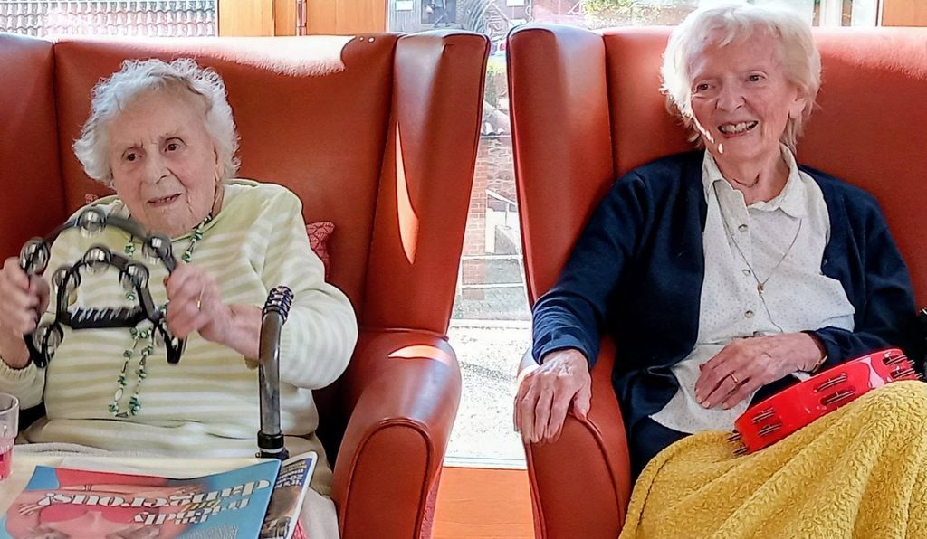 During visits, Andrew and Marko delight in seeing two elderly ladies seated in orange chairs, both smiling and holding tambourines—one wears a striped jumper and holds a newspaper, the other wears a white blouse with a yellow skirt.