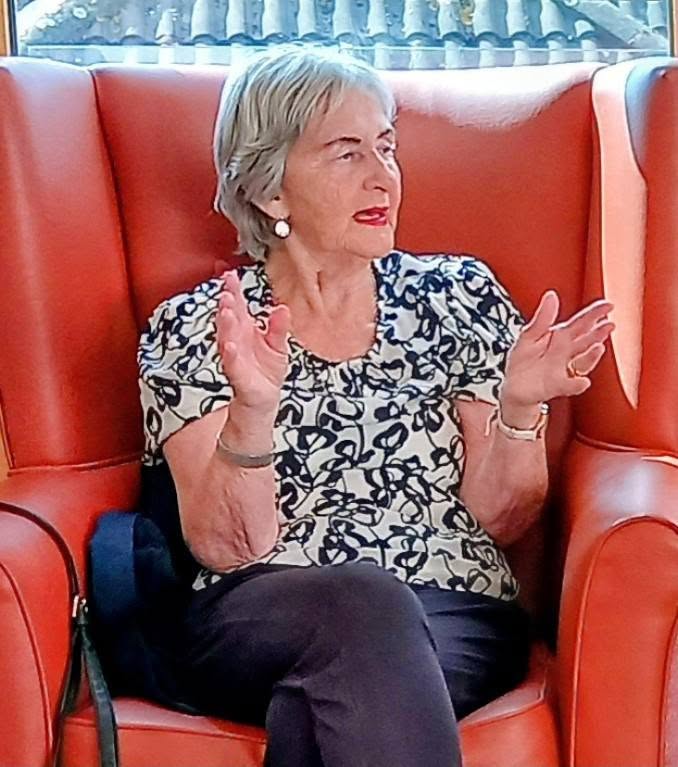 An older woman with grey hair sits in an orange chair, gesturing as she speaks during Marko’s visit. She is wearing a patterned blouse, dark trousers, and earrings.
