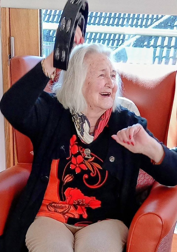 An elderly woman with white hair sits in an orange chair, smiling and raising a tambourine. Sunlight streams behind as she enjoys visits from Andrew and Marko, dressed in a black jacket, floral shirt and light trousers.