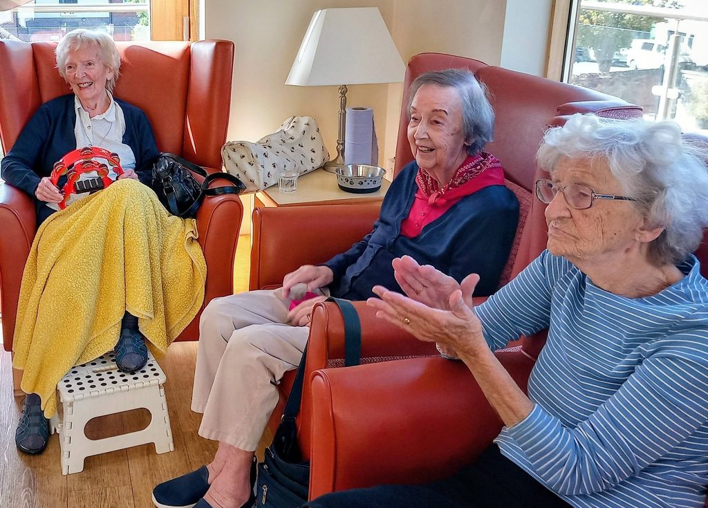Three elderly women sit in red armchairs in a well-lit room during Andrew and Marko’s visits—one holds a yellow blanket, one claps, and another smiles. A lamp and some bags are on the small table behind them.