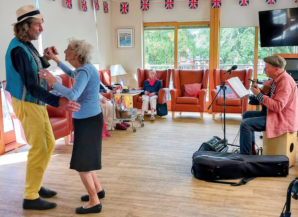 During Andrew’s visit, an elderly man and woman dance as a musician plays guitar in a bright room adorned with Union Jack flags. Other pensioners sit in armchairs, watching the cheerful scene.