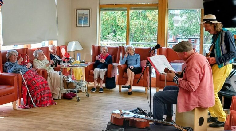 A group of older adults sit in armchairs watching musicians Andrew and Marko perform in a bright room. Musical instruments lie scattered on the floor, with large windows showing greenery outside during their visits.