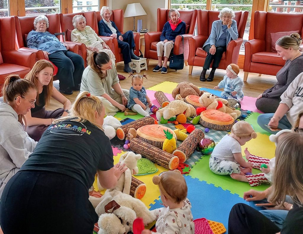 Babies and adults sit together on colourful mats with toys in the centre of a bright room, enjoying Baby Boogie dance classes, whilst older adults observe from armchairs by the wall.