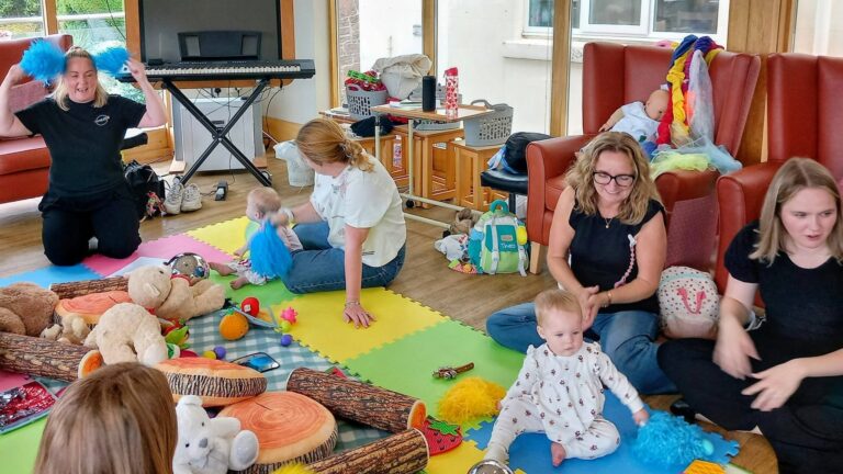The group seems to be attending a Baby Boogie baby dance class, with adults and babies sitting on colourful foam mats, toys and musical props scattered about, and a keyboard visible in the background.