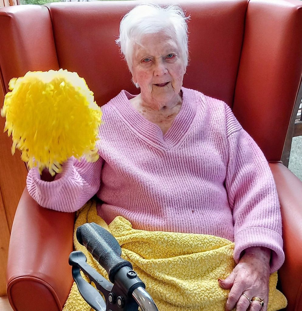 Elderly woman with white hair sits in a red armchair, wearing a pink jumper and yellow blanket, holding a yellow pom-pom—perhaps recalling dance sessions or watching nearby Baby Boogie classes. A walking frame stands beside her.
