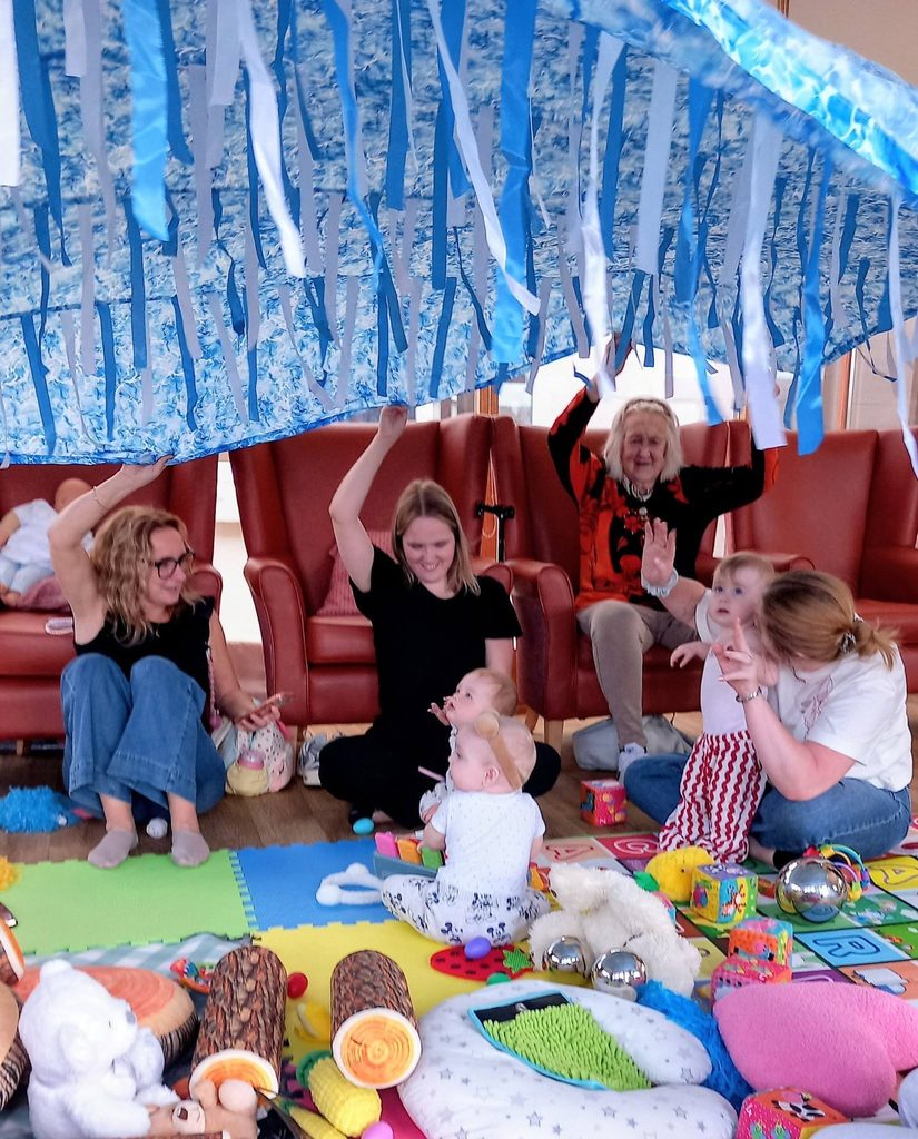 Adults and babies sit on a playmat with toys, gazing up at a blue and white canopy—ideal for Baby Boogie or baby dance classes to inspire movement and fun beneath cheerful streamers.