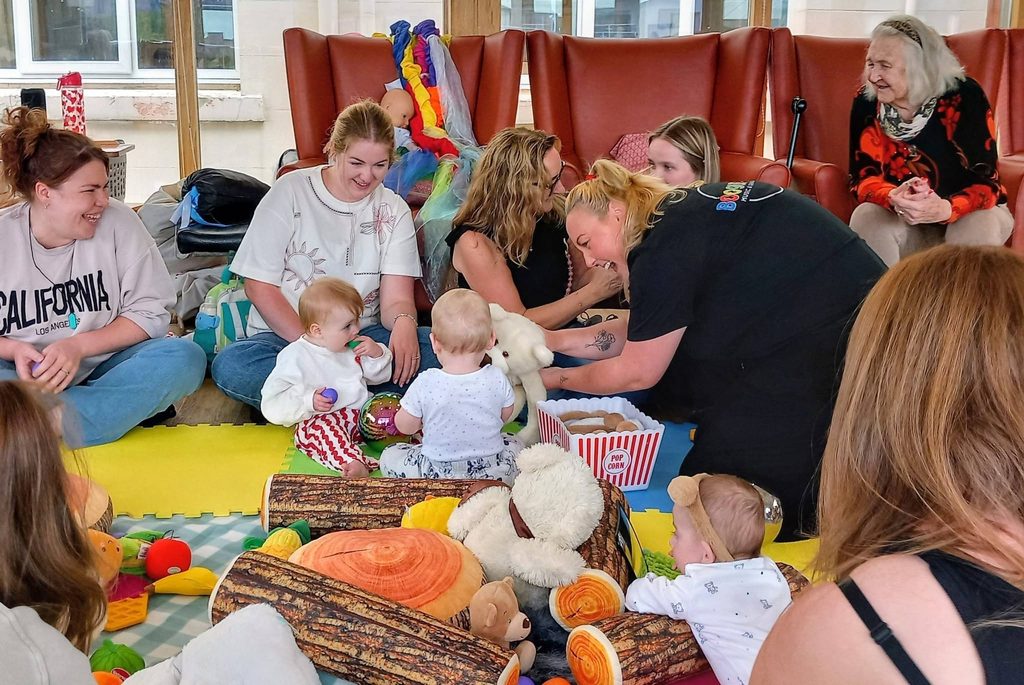 A group of women and their babies enjoy playtime on a mat strewn with toys, chatting about baby dance classes. An elderly lady sits in the background, smiling warmly at the lively group.