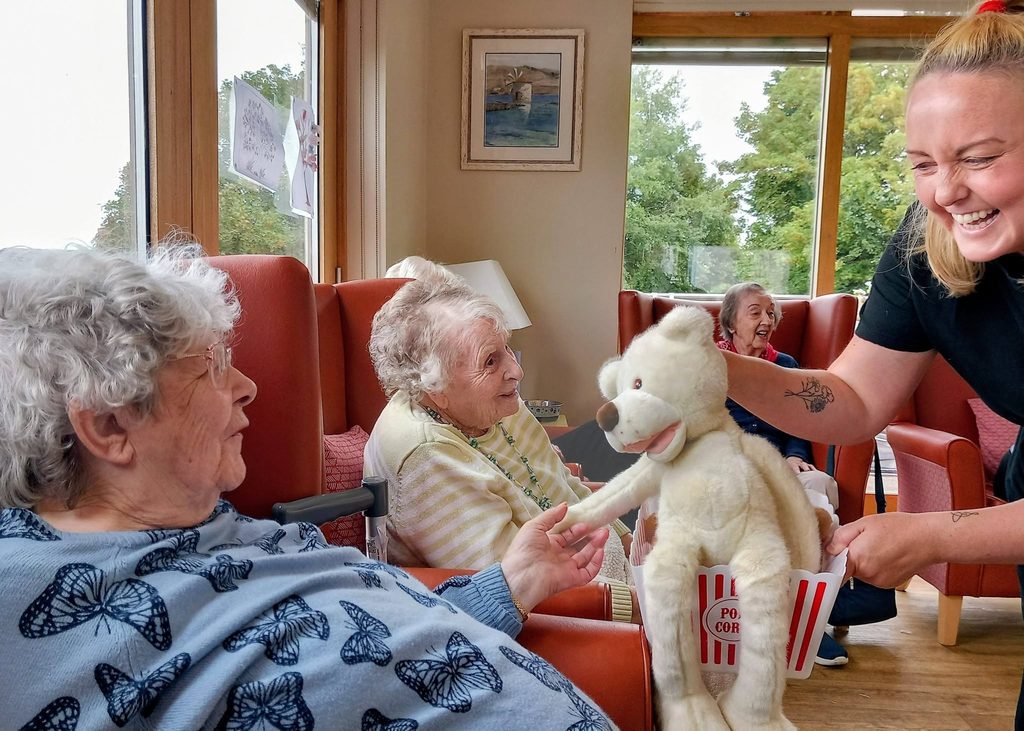 A carer amuses elderly ladies in a care home, holding a white bear puppet and popcorn bucket, evoking Baby Boogie dance session smiles, as greenery glows through large windows.