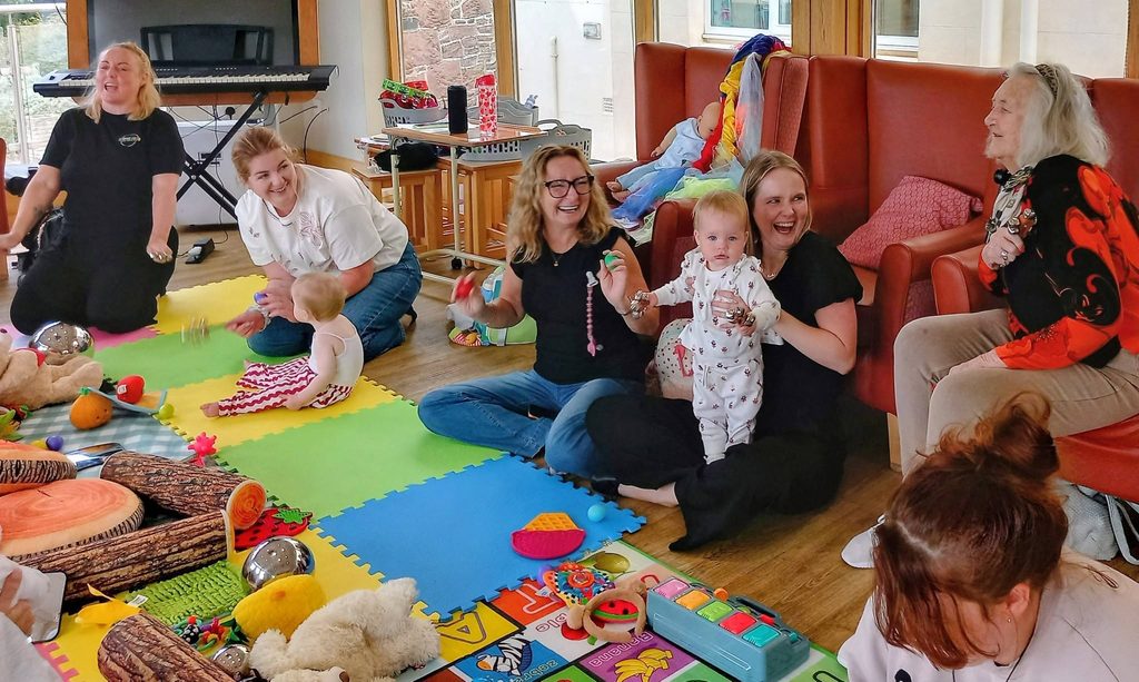 Adults and children enjoy Baby Boogie on colourful foam mats in a bright room. Some hold babies, others interact and smile, with toys scattered about during lively children’s dance sessions.