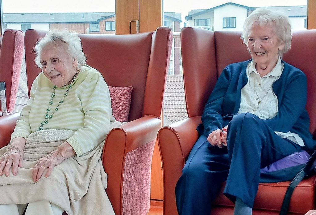 Two elderly women sit in high-backed chairs indoors, smiling and at ease as sunlight pours in. The room hosts lively Baby Boogie sessions with youngsters, whilst city rooftops are visible through the large windows behind them.