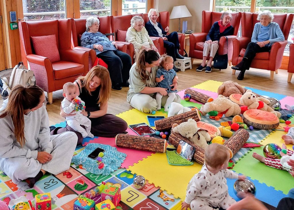 Babies play with toys on colourful mats as adults watch and smile in a bright room, ready for Baby Boogie or baby dance classes to begin.