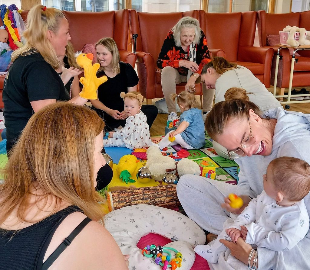 Women and babies sit together on the floor in a brightly lit room, surrounded by toys—some taking part in Baby Boogie dance sessions, while others watch and smile. A woman in a red and black top sits in the background.