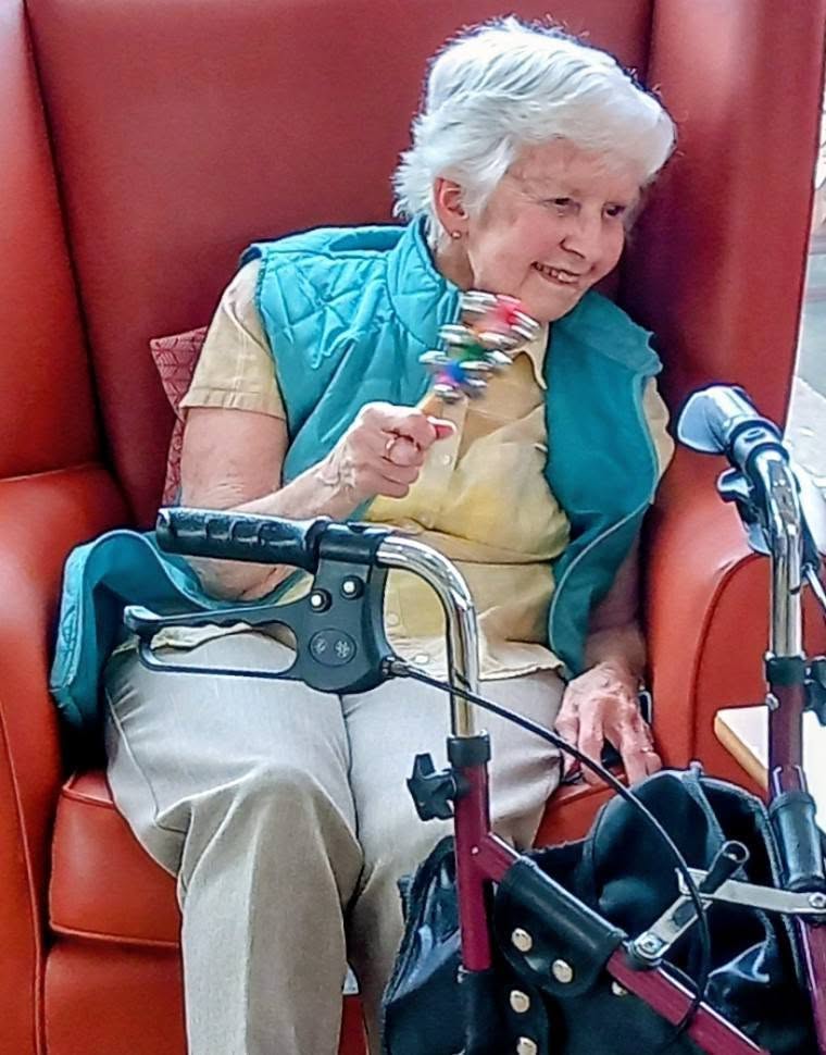 An elderly woman sits in a red armchair holding a colourful rattle, with a walking frame and black bag close by, as she enjoys the lively Baby Boogie sessions in the new block.