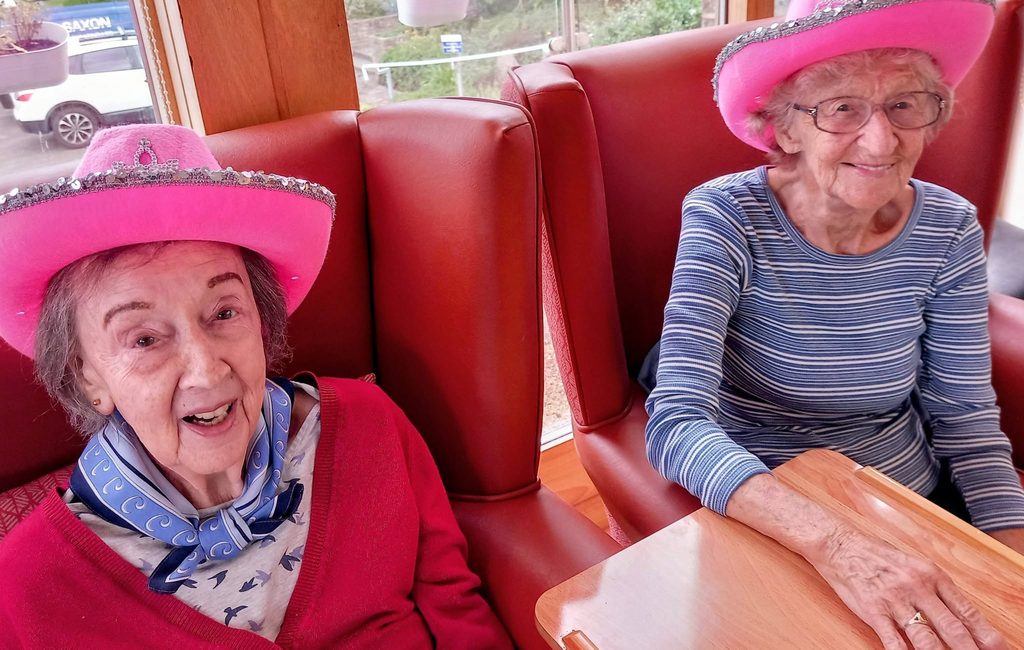 Two elderly women sit together in a restaurant booth, both smiling and wearing bright pink cowboy hats for Wild West Week. One wears a striped top and spectacles, while the other has a scarf tied round her neck.
