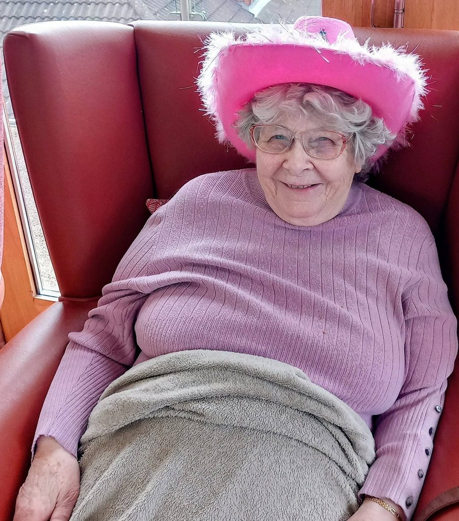 Elderly woman seated in a red chair, smiling, wearing spectacles, a pink feathered hat for Wild West Week, and a purple jumper, with a beige rug draped over her lap.