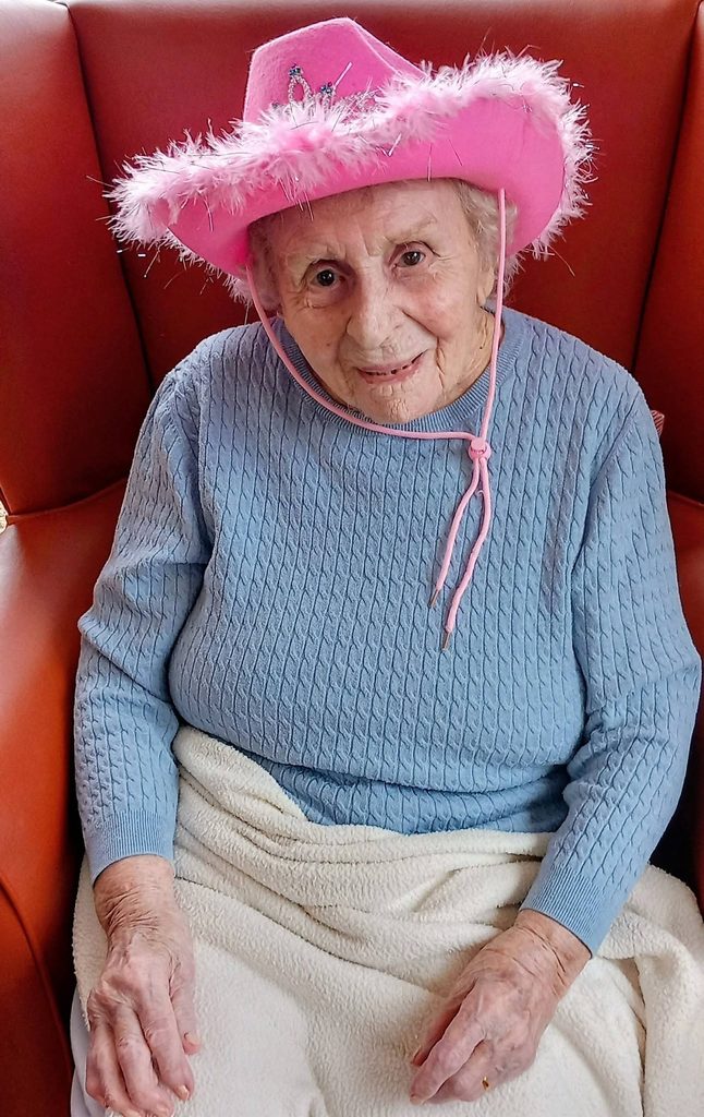 Elderly woman sits in an orange chair, snug beneath her blanket, sporting a pink cowboy hat trimmed with feathers—set for Wild West Week with proper Boogie Baby flair!.