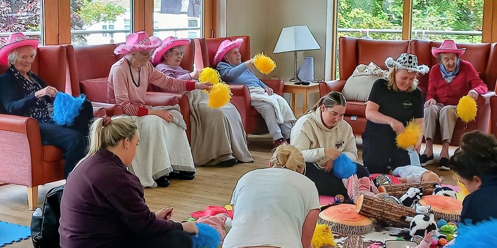 A group of elderly women in pink hats sit on sofas waving yellow pom-poms as Boogie Baby begins, whilst younger adults and babies play with colourful items in a bright room with large windows.