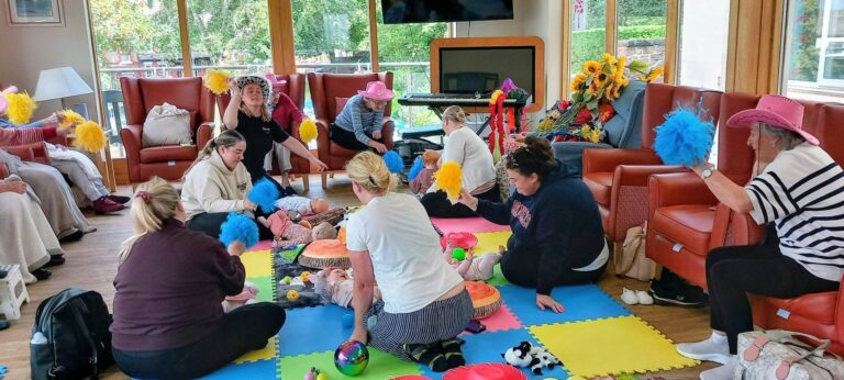 A group of adults and babies sit on colourful foam mats in a bright room, shaking pom-poms for Boogie Baby’s Wild West Week. Toys and sunflowers add cheer as everyone joins in the fun!.