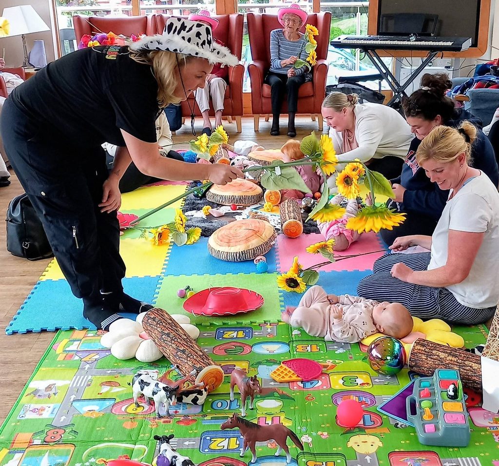 During Boogie Baby’s Wild West Week, adults and babies gather on colourful mats with toys, sunflowers, and animal props as a leader in a cow-print hat encourages everyone to join in playful activities.