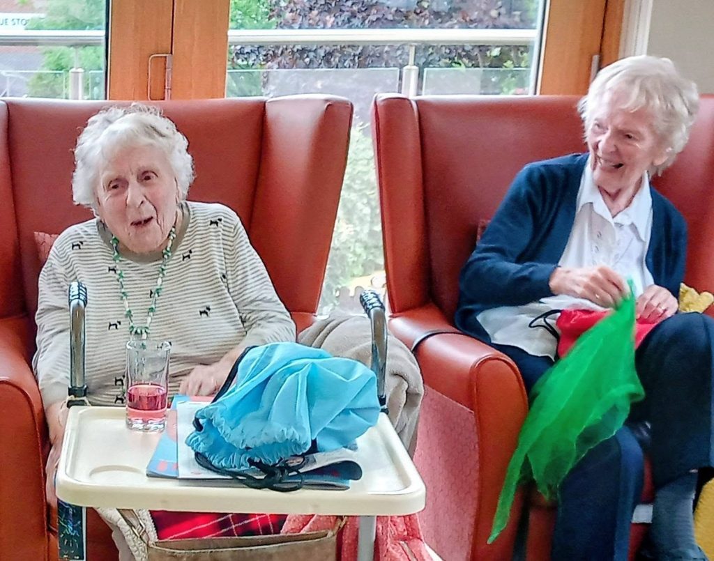 Two elderly women sit in red chairs by a window. One has a drink on a tray, whilst the other smiles, holding colourful fabric—perhaps reminiscing about dancing or planning to sing together.