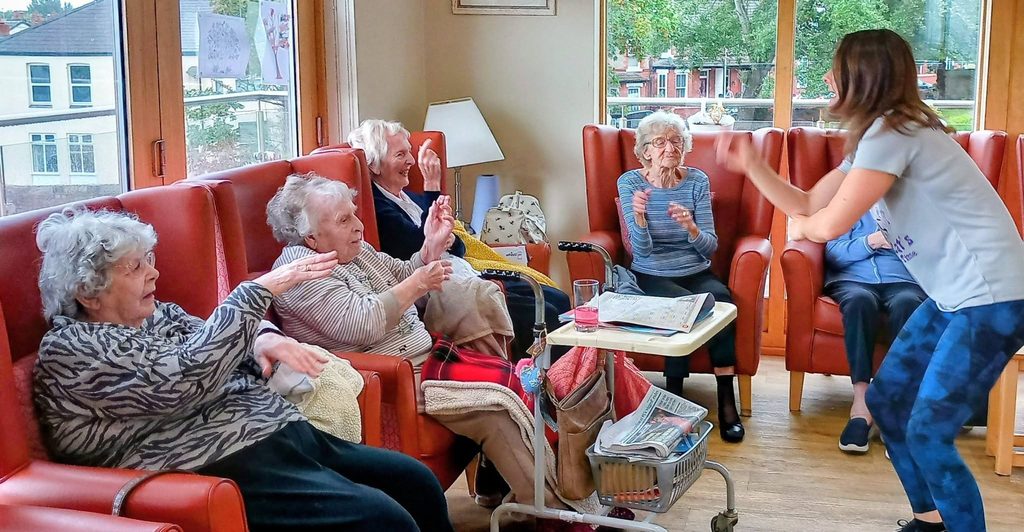 A group of elderly women sit in armchairs, watching and engaging as a woman stands to lead a lively quiz. Magazines and drinks are on a nearby table. Large windows overlook houses and gardens outside.