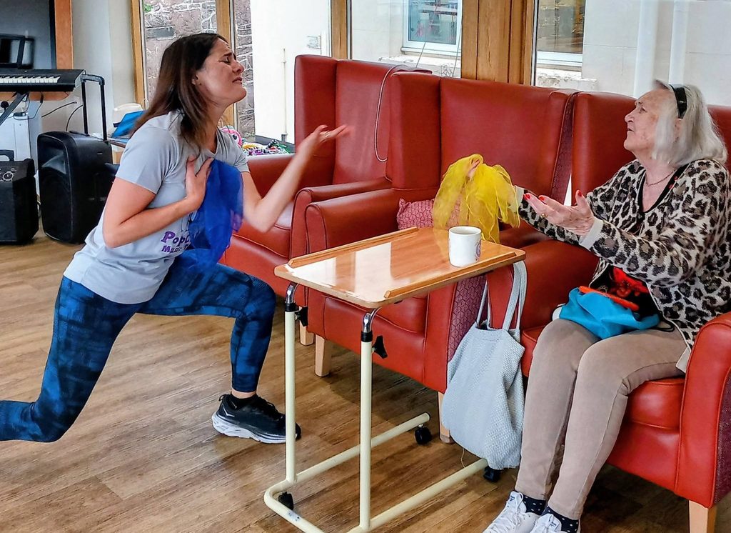 A woman kneels, gesturing expressively as if inviting the older woman on the red sofa to sing or quiz her; a cup and bags rest on the table between them in a relaxed indoor setting.
