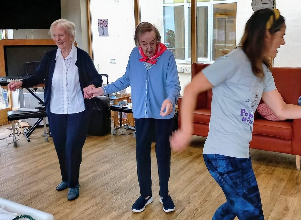 Two elderly women hold hands and smile as they join a group dance activity with a younger woman in a bright indoor room featuring large windows.