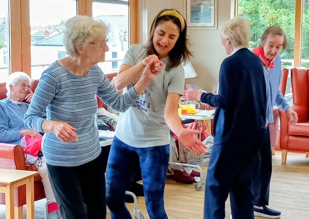 A group of older adults and a younger woman dance and sing together in a bright, communal lounge with large windows and comfy seating.