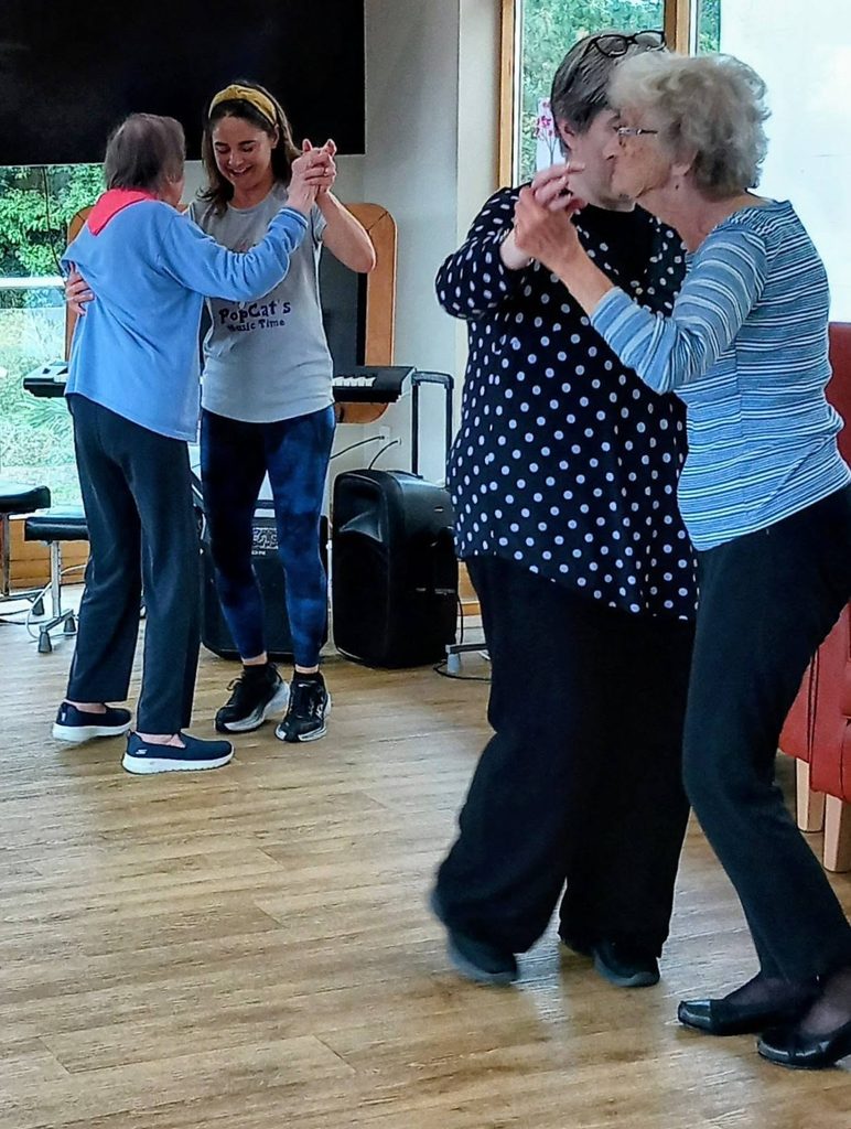 Two pairs of women dance together in a bright room with wooden flooring; a piano and speaker can be seen, creating the ideal atmosphere to sing or enjoy a lively quiz.