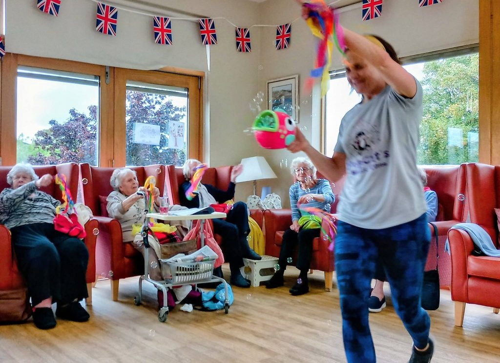 A group of older adults sit in armchairs in a decorated room as a woman at the front blows bubbles with a toy, involving everyone in a fun activity. Union Flag bunting hangs overhead as they get ready for a singalong or quiz.