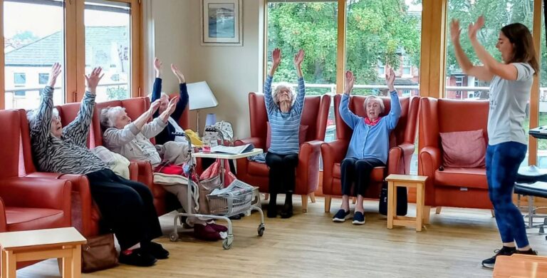 A group of elderly people seated in chairs raise their arms, following a woman leading an energetic exercise and dance session in a bright room with large windows.