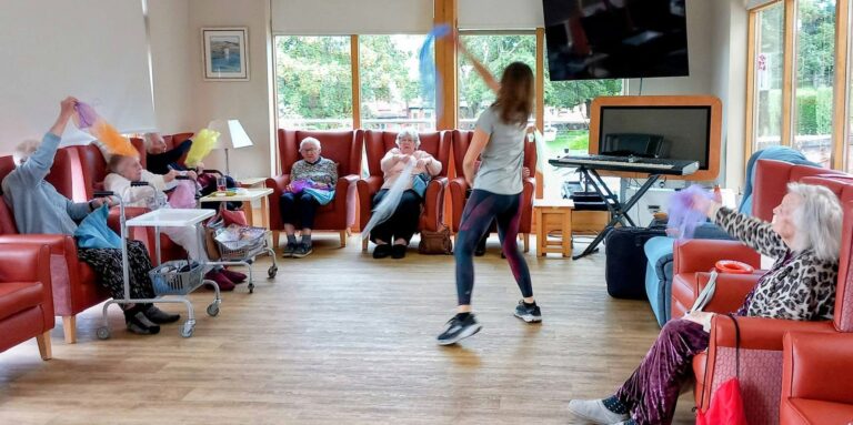 Caroline hosts a splendid afternoon as older people relax on red chairs by large windows, waving scarves together before forming a conga line through the lounge.