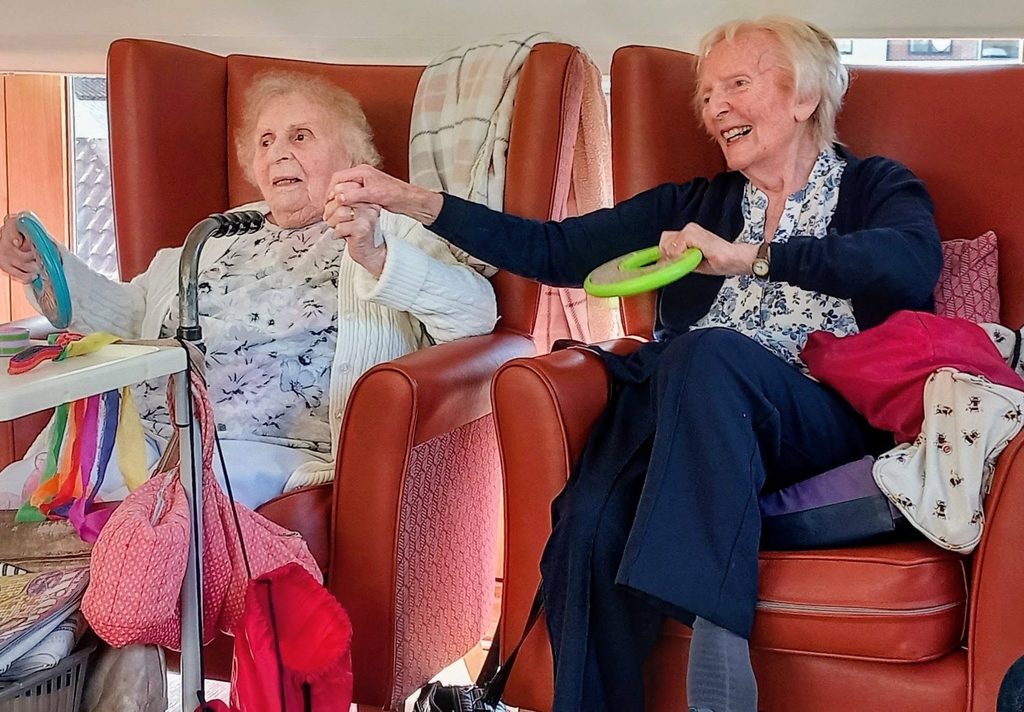 Two elderly women sit in armchairs, smiling and holding colourful plastic rings, enjoying a fabulous afternoon together. Personal items and fabrics lie about, adding a cosy atmosphere to the cheerful scene.