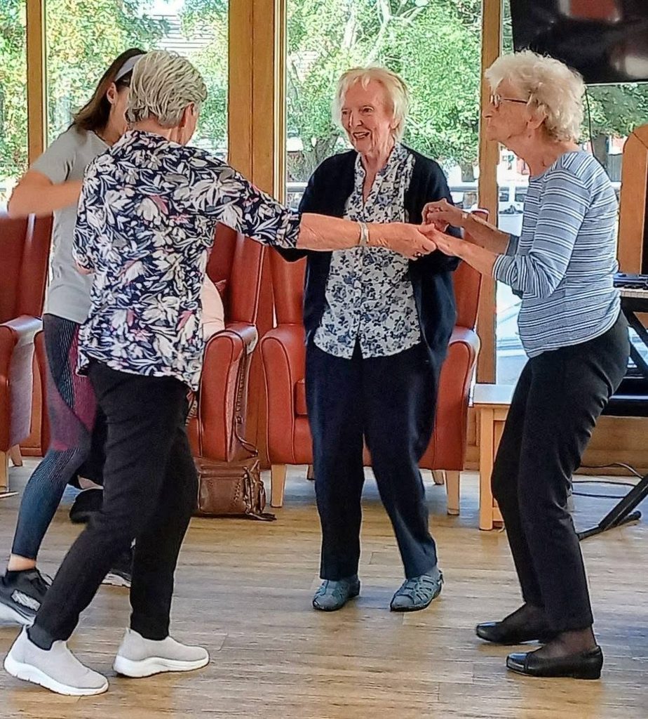 Three elderly women form a cheerful conga line, holding hands and dancing in the bright lounge with large windows. Caroline and another person stand nearby, both smiling and sharing in the enjoyment.
