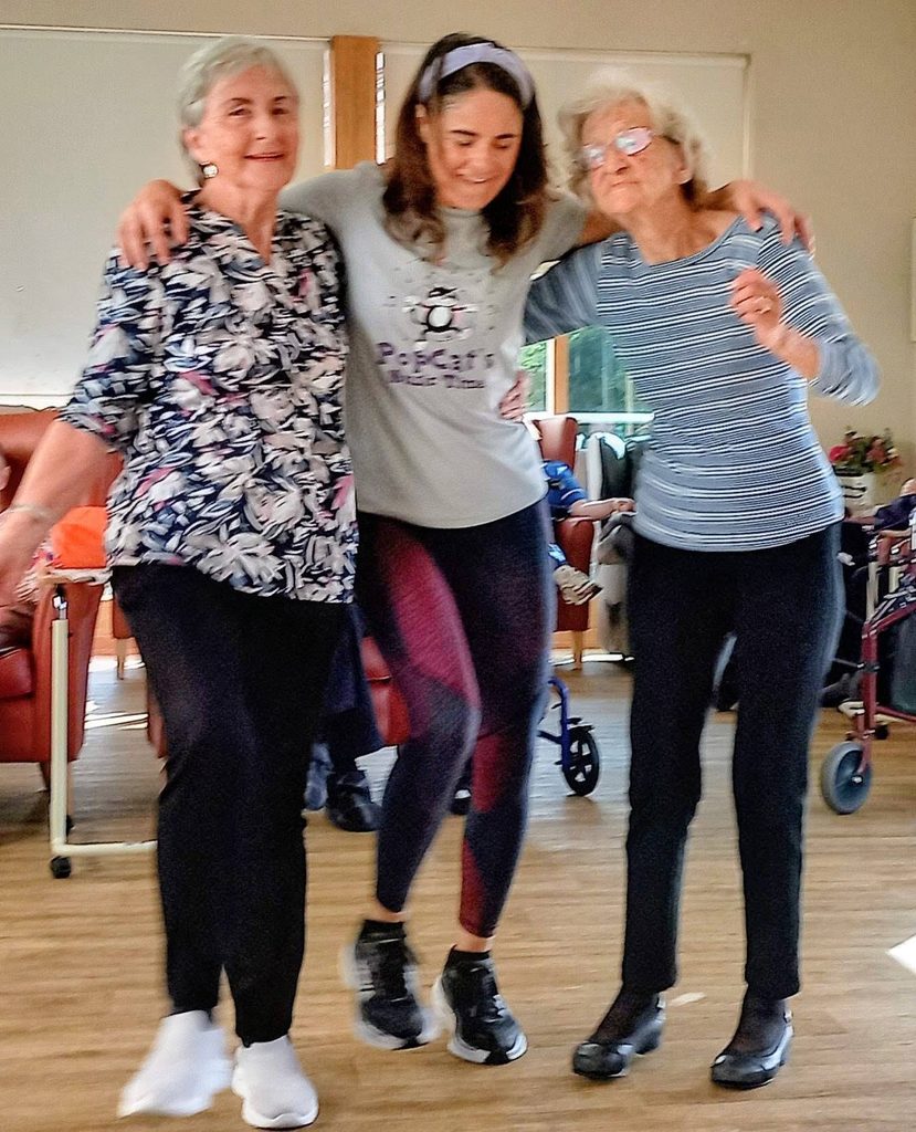 Three women, including Caroline, stand indoors with arms round each other, smiling and playfully posing—it’s a Conga Through the Lounge moment on a splendid afternoon in their shared living space.