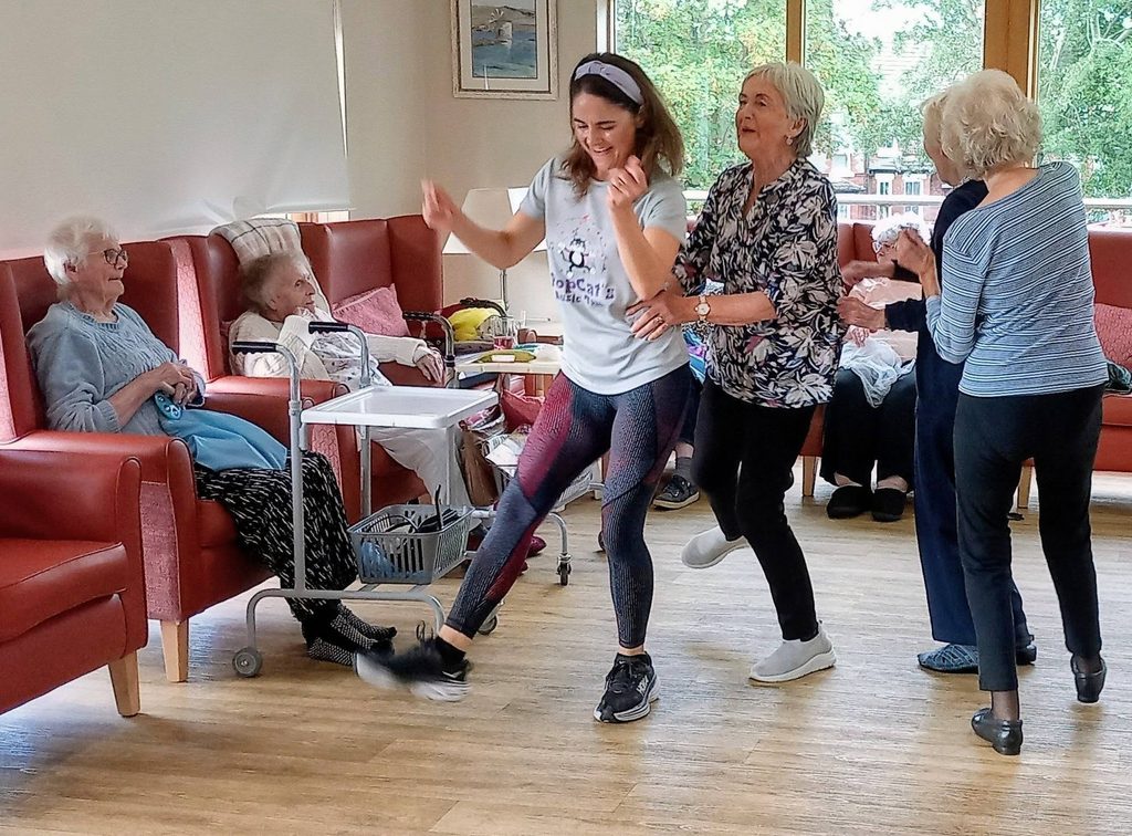 Caroline leads a conga through the lounge as elderly ladies and a younger woman dance in the bright room, whilst two others enjoy this lovely afternoon from red chairs next to a white walking frame and small table.