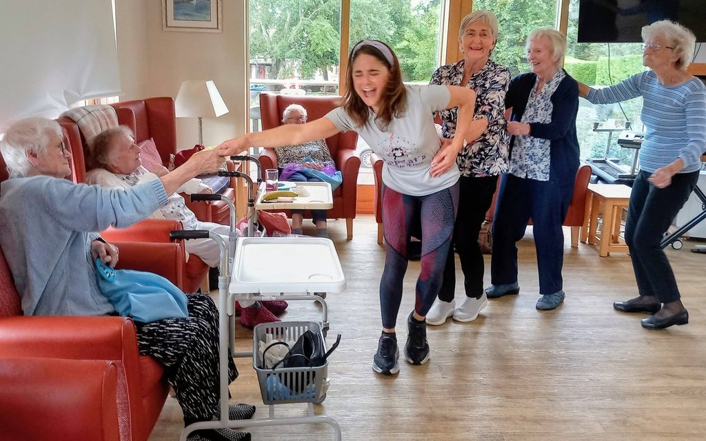 Caroline leads a conga through the lounge as a group of elderly ladies and a younger woman laugh, hold hands, and enjoy a marvellous afternoon together in the care home lounge.