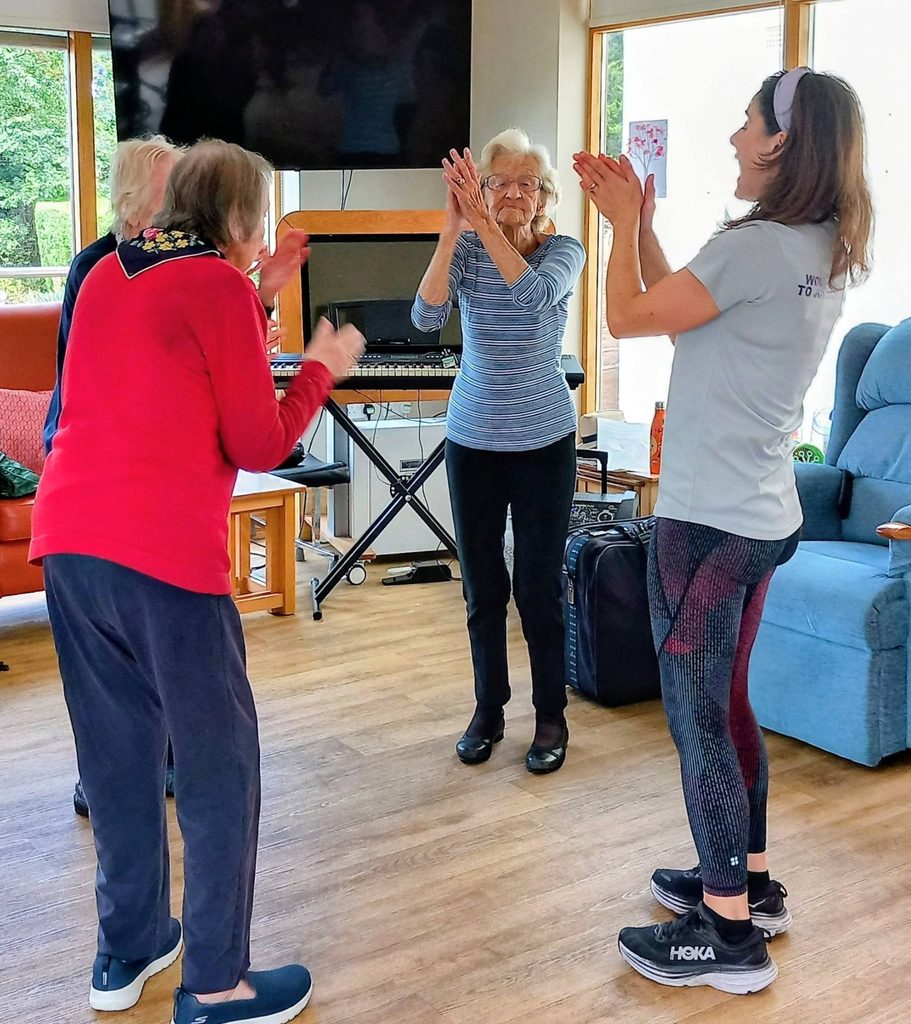 Four women stand in a bright lounge, clapping and smiling together during what seems to be a group activity or conga exercise session.