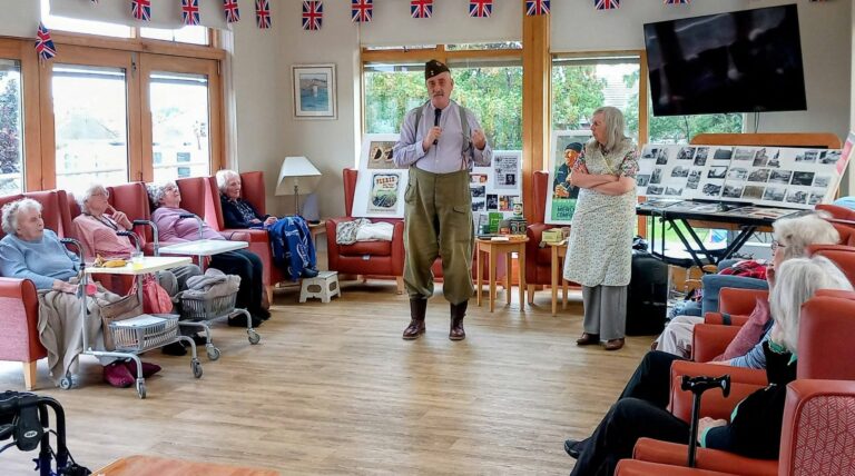 A man in historical costume addresses elderly people seated in a decorated room, whilst a woman stands nearby and historic photographs are displayed on boards.
